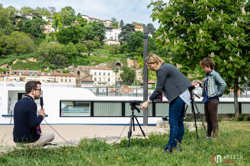 Interview en extérieur réalisé par Deborah Donnier au bord de la rivière pour le documentaire 'Think WP', Aurélien Denis au micro face à une caméra sur trépied, ville en arrière-plan