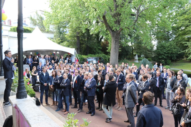 Discours en plein air devant une foule dense de professionnels dans un parc arboré, vue en hauteur