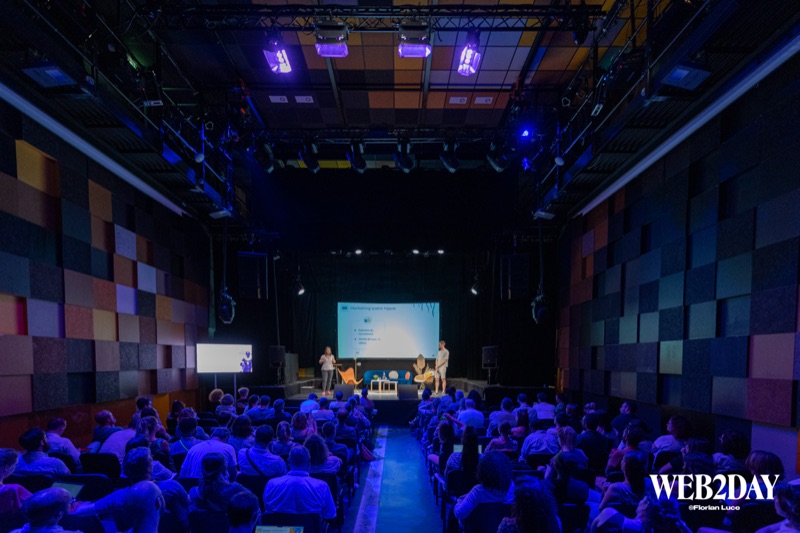 Vue grand angle d'une salle de conférence avec public assis face à la scène, éclairage bleu et panneaux acoustiques colorés
