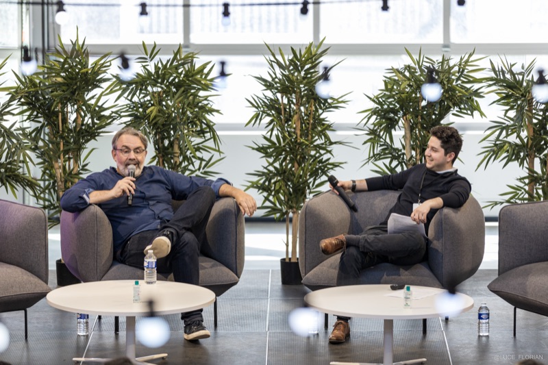 Deux intervenants échangeant en fauteuils lors d'une conférence, plantes et baies vitrées en arrière-plan