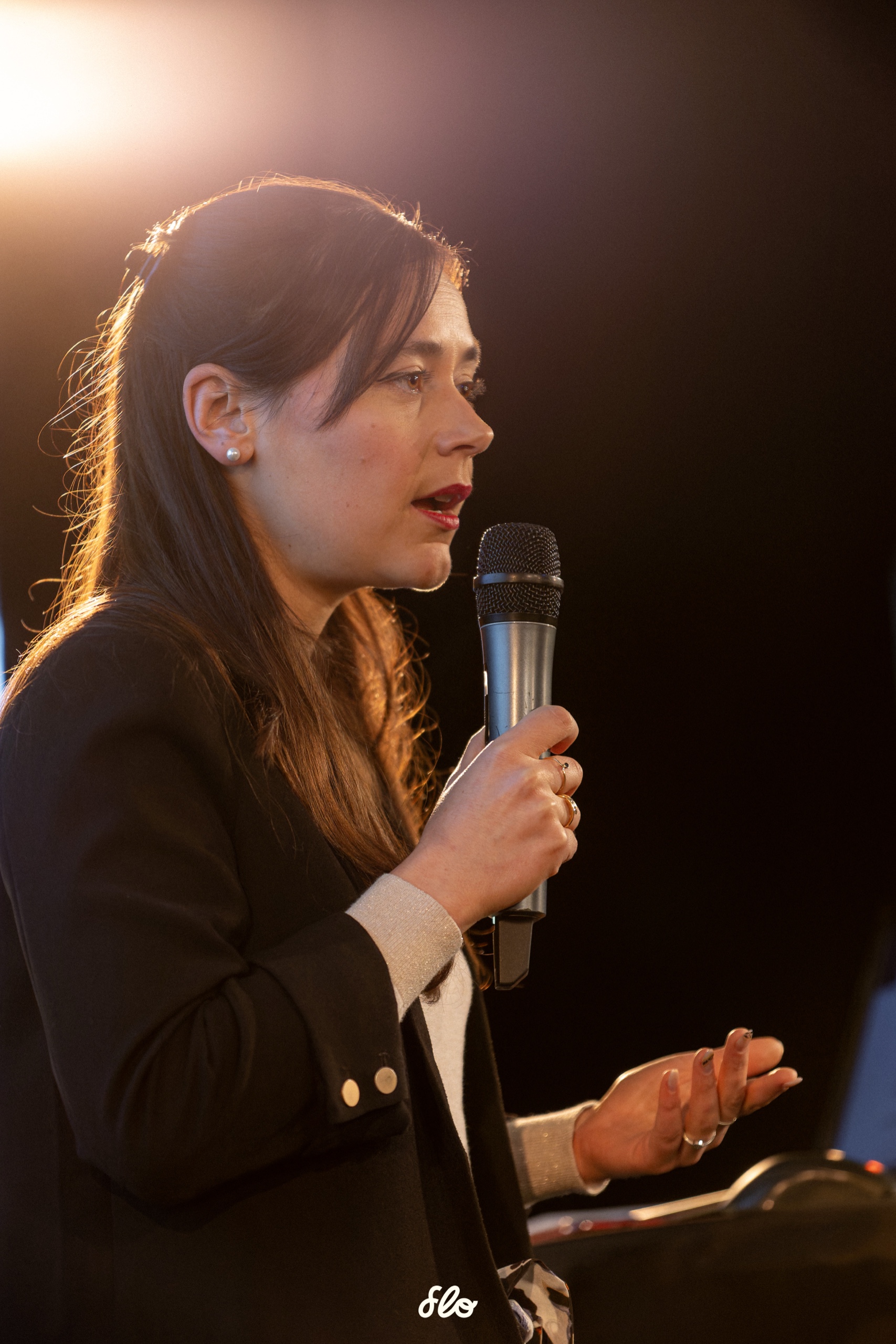 Maître Graziella Dode, avocate dédiée au droit des animaux, de profil au micro lors d'une conférence, contre-jour doré sur fond sombre