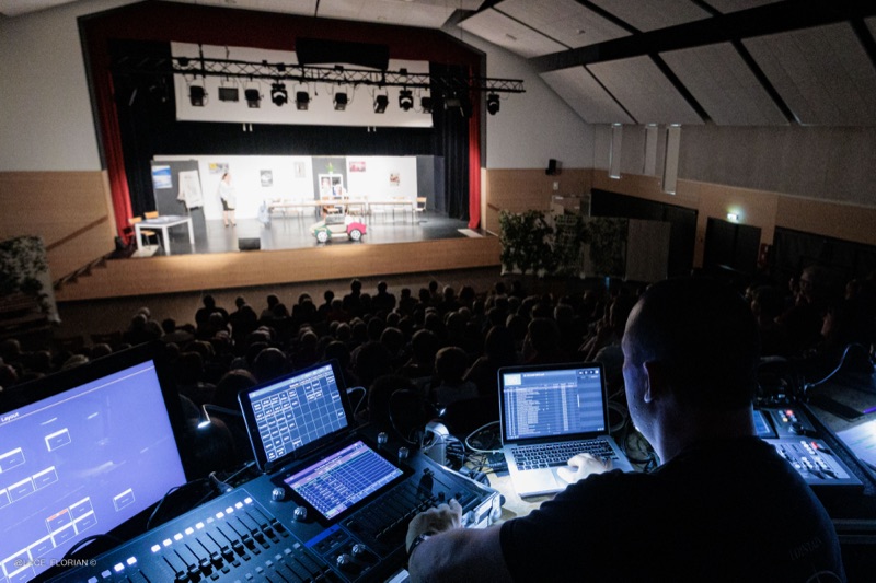 Régisseur technique devant ses consoles et écrans, vue plongeante sur la salle de spectacle