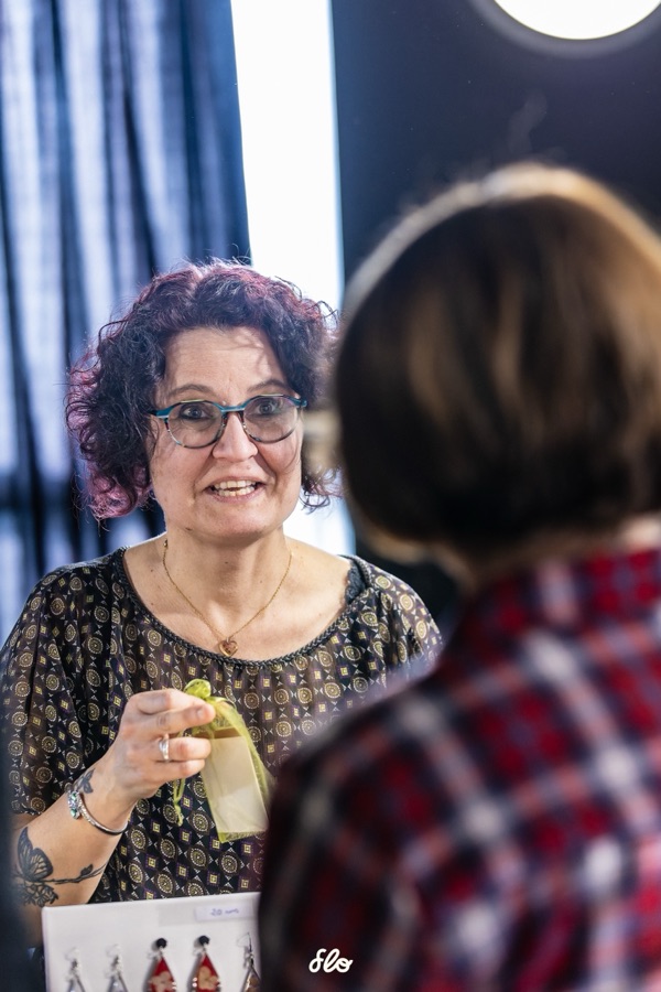 Exposante souriante présentant un bijou artisanal à une visiteuse en chapeau