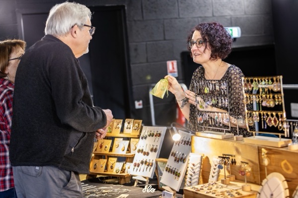 Stand de bijoux artisanaux avec présentoirs en bois, exposante échangeant avec un couple de visiteurs