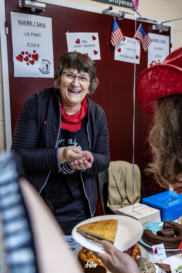 Bénévole souriante servant une part de galette derrière le comptoir décoré de cœurs